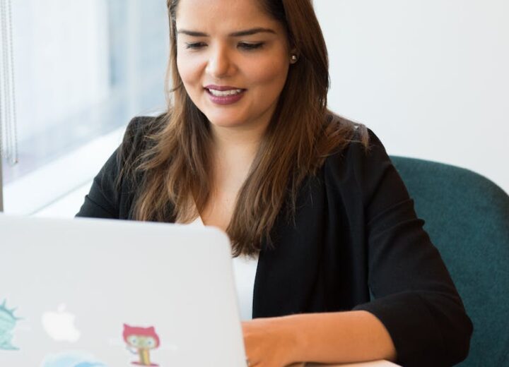 Smiling woman in black blazer working on laptop with open notebook on desk in a modern office setting.