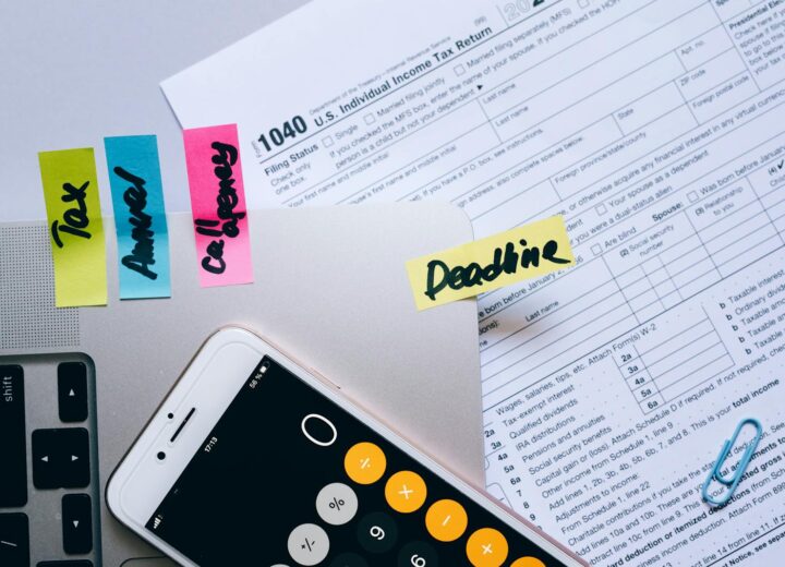 Office desk flat lay showing tax documents, calculator app on smartphone, sticky notes, and paperclips.
